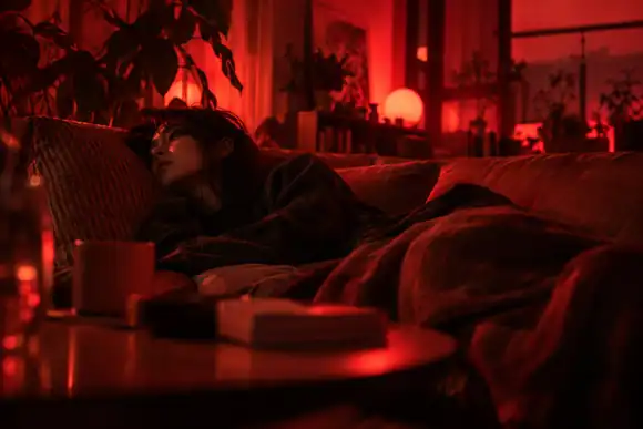 Une jeune personne fatiguée allongée sur un canapé dans une pièce sombre baignée de lumière rouge